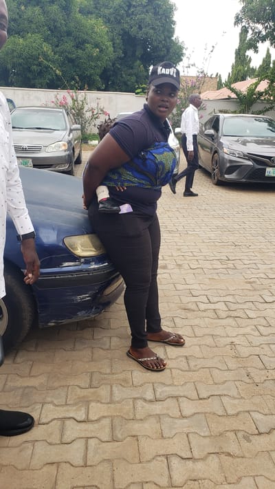 In Unguwar Shanu, Nigeria, a woman wearing a black baseball cap that says "KADVS" and black leggings is leaning against the hood of a damaged blue car. She is holding a baby wrapped in a blue and green patterned cloth, with the baby's legs dangling. The baby is wearing black shoes. A man in a white shirt and black pants is walking away from the scene in the background, past a grey car. Another person, mostly out of frame on the left, is wearing a white patterned shirt. The ground is paved with interlocking bricks, and there are trees and a building in the background. The blue car has visible damage on its front bumper.