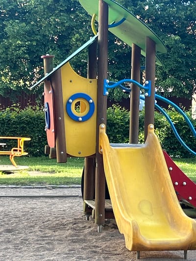 The image shows a colorful playground structure with wooden posts and vibrant yellow and red slide sections, set against a backdrop of lush green trees and a wooden fence. The playground features a yellow house-like section with a circular blue and yellow window. To the left, a yellow and orange piece of playground equipment, possibly a merry-go-round or a spinning seat, is partially visible. The ground is covered in sand and grass. This playground is located at Norra Bergens Koloniområde in Helsingborg, Sweden. There are no people or animals visible in the image. The scene appears to be an outdoor public space, likely a park or recreational area, bathed in daylight.