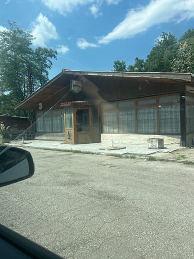 The image shows a low-rise, wooden building with a pitched roof, possibly a restaurant or shop, situated on a paved area in the city of Dolna Vasilitsa, Bulgaria. The building has large windows covered by patterned curtains and a small, enclosed entrance. A sign reading "Kamenitza" is visible on the facade, indicating a brewery or beverage brand. To the left of the building, there are trees and a tall metal structure, possibly a pylon or antenna. A car's side mirror is partially visible in the foreground, suggesting the photo was taken from a vehicle. The ground is a cracked asphalt surface, and there are some plants growing along the edge of the pavement. The sky is blue with scattered white clouds.