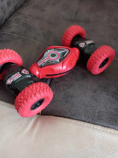 The image shows a red and black remote-controlled stunt car with large red wheels, positioned on a textured brown leather surface. The car has a sleek, futuristic design with black accents and red detailing on its body. It appears to be lying on a sofa or armchair. The background is a close-up of the textured leather. There is no indication of the location being in Кош-Дөбө, Kyrgyzstan; this information is extraneous to the visual content of the image.