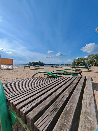 The image depicts a sandy beach with several empty green metal-framed, wooden slatted lounge chairs. The lounge chairs are arranged in rows on the sand, facing a body of water. The water appears to be a lake or a large river, with small waves and a light breeze. In the distance, there are several small, tree-covered islands and a line of trees along the shore. The sky is blue with scattered white clouds. On the left side of the image, there is an empty orange signpost with a blank rectangular sign. The scene is likely a public beach area, possibly in the vicinity of Zhdanovichi, Belarus, a location known for its recreational areas and proximity to water bodies. The overall impression is one of a peaceful, undeveloped natural setting, ready for visitors.