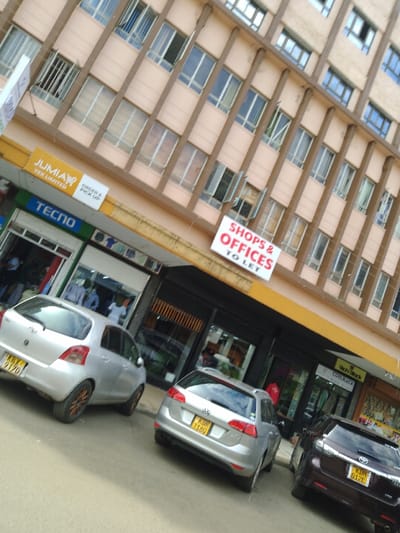 The image shows a street-level view of a commercial building located at the coordinates -1.453, 37.007 in Athi River, Kenya. The building is a multi-story structure with a tan facade and large rectangular windows. The building has a sign advertising "Shops & Offices To Let". There are three cars parked in front of the building. The car closest to the viewer is a silver hatchback with the license plate KBY 8170. The second car is a silver wagon with the license plate KDB 2180. The third car is a maroon sedan with the license plate KDH 012E. In the background, a pedestrian is visible walking on the sidewalk. The shop front closest to the viewer is a retail store with "TECNO" written on the awning. The shop on the far right is a store called "Cool Black" with a dark sign.