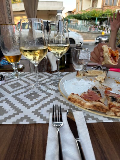 A table is set for a meal, with a half-eaten pizza on a plate, several glasses of white wine, a fork, and a knife. A person's hand is visible in the upper left, holding a piece of the pizza. The table is covered with a patterned tablecloth and rests on a wooden surface. In the background, a building with brickwork and windows can be seen, along with greenery and a white vehicle. The scene appears to be outdoors, possibly at a restaurant, with natural light suggesting it is daytime. The overall impression is of a casual meal shared among people.