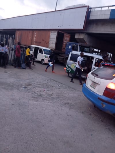 The image depicts a traffic scene in Port Harcourt, Nigeria, likely on an elevated road or bridge. In the background, a large container truck has partially fallen or tilted precariously, with its cargo container resting at an angle against the overhead structure. Below this, a blue truck is visible. In the foreground, several people are standing on or near the road, some appearing to be passengers or onlookers. A white mini-bus with a cartoon decal on its side is parked with its doors open, and a man in a white t-shirt is walking away from it carrying a bag. To the right, the rear of a white and blue car is visible, with its taillights illuminated. The road surface appears to be asphalt or concrete, and there is some debris scattered on the ground. The sky is overcast.