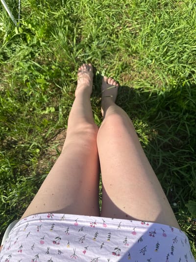 An overhead shot shows a person's legs and feet in green grass. The person is wearing a white garment with a floral print. The sunlight casts shadows on the grass, suggesting it is daytime. The location is implied to be a park or backyard in Mokino, Russia. The scene is peaceful and natural, with no other people or objects visible.