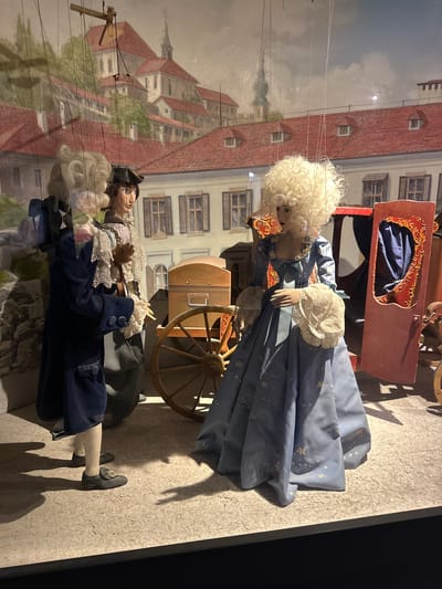 This image shows a display of marionettes dressed in historical costumes, set against a backdrop depicting a cityscape with buildings and a church tower, suggestive of Salzburg, Austria. The puppets are arranged in a scene that includes a red carriage with ornate gold detailing and a wooden cart with a large wheel. Three primary marionette figures are visible: one on the left, wearing a dark blue coat and a white wig; a second in the center, in a white ruffled shirt, brown vest, and a black hat; and a third on the right, a female figure with a large, curly blonde wig, wearing a light blue, floor-length gown adorned with floral patterns. The female puppet is holding a white ruffled accessory in her hand. The figures are suspended by strings, indicating they are part of a puppet show or exhibition. The lighting casts shadows on the textured ground, enhancing the theatrical presentation.