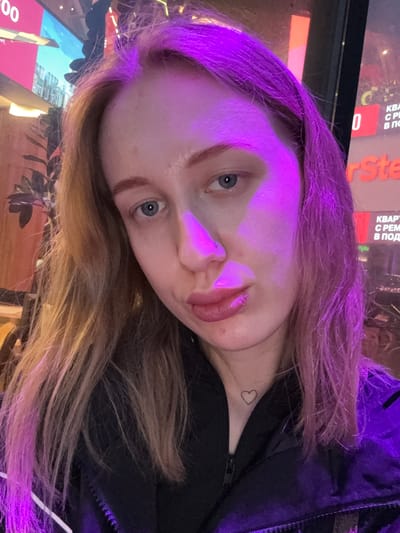 A woman with light skin and blue eyes is depicted in a close-up shot. The image is rotated counter-clockwise, casting a purple light over the upper portion of her face and hair. The woman has reddish-blonde hair that fades into lighter shades towards the ends. A small heart tattoo is visible on her neck. She is wearing a black jacket. In the background, blurred text in Russian can be seen, including the name "Ste" from a possible brand or logo. The location is likely Ol'ginskaya, Russia, based on the Russian text in the background.