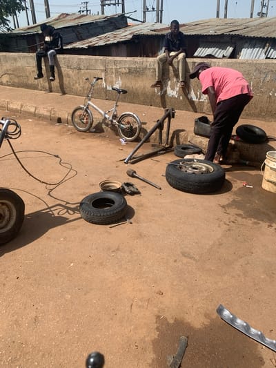 In Katon Rikkos, Nigeria, two young men are seated on a concrete wall, one in a black hoodie and the other in a blue t-shirt. A third man, wearing a pink t-shirt and a dark cap, is bent over near a pile of tires and a black plastic container. A small white bicycle is parked on the ground nearby. The scene appears to be an outdoor workshop or repair area, with various tire-related tools and spare tires scattered on the dusty ground. The background shows a row of corrugated metal roofs, suggesting a bustling urban or semi-urban environment. The overall impression is one of daily life and work in a Nigerian community.