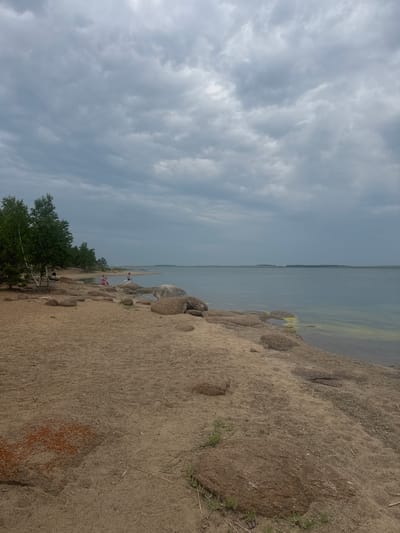 This is a horizontal, eye-level, outdoor, full shot of a sandy beach area with a body of water under a cloudy sky. The foreground is dominated by a sandy beach scattered with rocks of various sizes and some sparse green vegetation. In the middle ground, there are a few people scattered on the beach and near the water. To the left, there are several trees with green foliage. The background features a large body of water stretching to the horizon, with a distant treeline visible across the water. The sky is overcast with a thick layer of gray clouds. The scene appears to be a natural landscape, possibly a park or a recreational area, located in the city of Aiyrtau, Kazakhstan, known for its numerous lakes and natural beauty.