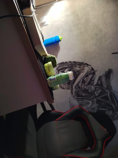 The image depicts a close-up, angled view of objects on and near a desk, with a portion of a black and red gaming chair visible in the foreground. On the desk, a bright blue water bottle with a green cap is lying down. Next to it, a green, textured object, possibly a sponge or stress ball, is visible, with a light green cylindrical object, possibly a travel-sized container of toothpaste or medication, lying on its side next to that. The background shows a gray floor with a large, detailed black and white graphic of mechanical components. The scene appears to be indoors, likely a home office or gaming setup. There are no people visible in the image, nor any indication of the specific location of "Крутые Выселки, Russia" beyond what is provided in the prompt.