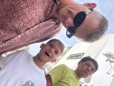 The image is a low-angle, wide-shot selfie taken outdoors. Three individuals are visible. In the foreground on the left is a young boy wearing a white t-shirt with a graphic design. To his right is an adult with short, dark hair, wearing a light green t-shirt and a gold necklace. In the background, looking down at the camera, is an adult wearing sunglasses with gold accents and a reddish-brown snakeskin-patterned shirt. Behind the individuals, a white building with vertical fins is visible. A sign on the building reads "ALE-HOP". To the right of the building, a large, stylized illustration of a cow is present on a white banner. The sky is overcast. The presence of the "ALE-HOP" store indicates the location is likely within a commercial area in Benidorm, Spain.