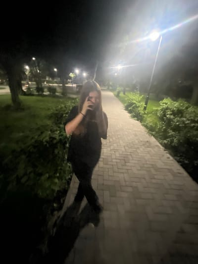 A young person with long brown hair, wearing a dark shirt and pants, stands on a brick pathway at night. The person is holding a phone to their face and appears to be taking a photo or video. Streetlights illuminate the pathway and surrounding greenery, casting shadows. The scene suggests an outdoor setting, possibly a park or walkway in the city of Bulgarovo, Bulgaria, given the context. The lighting is low, and the image has a somewhat grainy quality typical of nighttime photography.