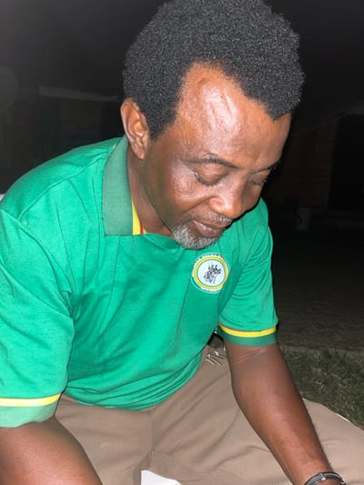 A man with short, dark hair and a trimmed gray beard is shown from the chest up, seated and leaning to his right. He wears a green polo shirt with yellow trim at the collar and sleeves and a circular patch with an image on the left breast. He also wears a dark bracelet on his right wrist. He seems to be relaxed and possibly sleeping. His eyes are closed and his expression is calm. The background is dark, with a hint of foliage visible in the upper right corner.
