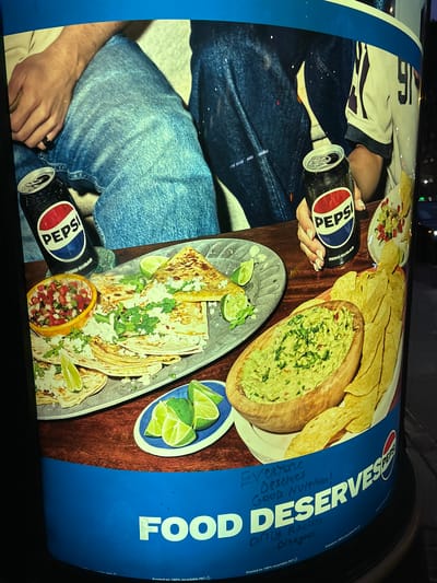 The image shows a cylindrical advertisement poster featuring a close-up view of a meal and Pepsi cans. The advertisement is predominantly blue and brown, with a prominent Pepsi logo.
In the foreground, a wooden bowl of guacamole is filled to the brim and garnished with chopped cilantro and red chili peppers. To the right of the guacamole are several tortilla chips, some broken and some whole, arranged in a fan-like pattern.
To the left of the guacamole, a metal plate holds a stack of quesadillas, cut into wedges. They are sprinkled with white cheese and garnished with cilantro. A small orange bowl of pico de gallo sits to the left of the quesadillas. Several lime wedges are scattered around the food items, and a few are on a small blue and white plate.
Two Pepsi cans are visible. One is slightly opened, with condensation on its surface, and features the red, white, and blue Pepsi logo. The other can, held by a hand with manicured fingernails, also displays the Pepsi logo and the text "ZERO SUGAR."
The background of the advertisement shows a portion of two individuals. One person is wearing blue denim jeans and their hand rests on their leg. Their other hand is holding the Pepsi can. The second individual, behind them, also appears to be wearing denim jeans.
Below the food imagery, the poster has a large blue section with white text that reads "FOOD DESERVES". There is also handwritten blue ink text above the printed text that says "Everyone Deserves Good Nutrition! Only Racists Disagree". The bottom of the poster includes printing information.
The image is a photograph of this advertisement poster, possibly on a public pillar or structure in an urban environment, given the lighting and the slight distortion of the cylindrical surface. The exact location in San Francisco, United States of America is not discernible from the visual content alone. The scene is well-lit, suggesting it was captured either during the day or under artificial street lighting at night.