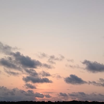 The image depicts a sunset sky with scattered gray clouds. The sky transitions from a pale gray at the top to a peachy-orange hue near the horizon, where the sun is partially obscured by clouds and the dark silhouette of a distant treeline. The location is identifiable as Badem, India, based on the image source.