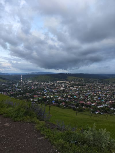 The media file depicts an aerial view of a town nestled in a valley, with rolling green hills and a hazy mountain range in the distance. The sky is predominantly covered by dark, dramatic clouds, suggesting an overcast day or perhaps an approaching storm. In the foreground, a grassy slope leads down to the town, with patches of wildflowers adding touches of color.
The town itself is a sprawling collection of houses, many with brightly colored roofs, interspersed with trees. A river or stream winds through the middle of the town, and a prominent, red-and-white striped smokestack stands tall on the outskirts, indicating an industrial presence. The architecture suggests a rural or semi-rural setting.
The time of day appears to be late afternoon or early evening, given the diffused light and the dark, brooding sky. The overall mood is somewhat somber due to the weather, but the vibrant rooftops and greenery offer a sense of life and resilience. The location is Sim, Russia, which aligns with the typical landscape of the region. There is no visible text in the image.