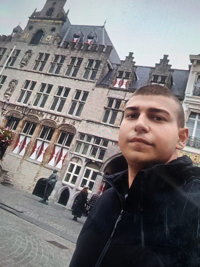 The image shows a young man with short hair taking a selfie in front of a historic building. He is wearing a black jacket. The building appears to be made of stone with multiple windows and decorative elements. In the background, other people can be seen walking in a plaza. There is a statue in the foreground. The sky is overcast. The location is identified as the city of Maasvlakte Rotterdam, Netherlands.