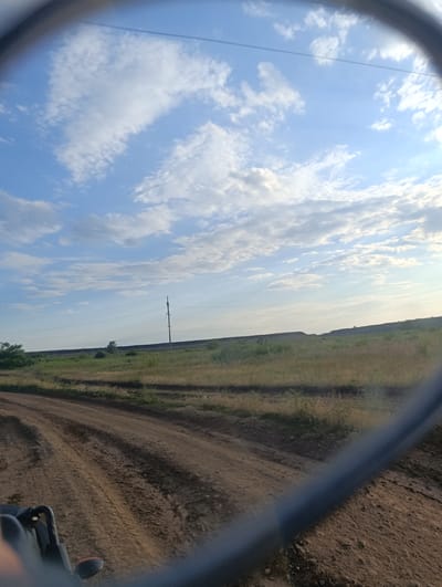 The image captures a rural landscape scene, viewed through a circular frame, possibly a mirror or a lens. A dirt road with tire tracks leads into the foreground. To the right of the road, dry grass and sparse vegetation cover the ground. In the distance, a gently rolling hill is visible, topped with dark material, possibly coal waste, characteristic of the area around Chystiakove, Ukraine, a region known for its coal mining industry. A single utility pole with power lines stands on the horizon. The sky above is a bright blue with scattered white clouds. The bottom portion of the image shows a blurred, dark-colored object, likely part of a vehicle.