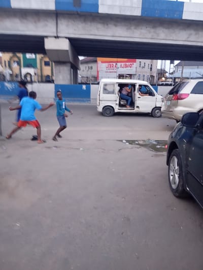 The image depicts a street scene in Port Harcourt, Nigeria, with an elevated highway overhead. In the foreground, the tarmac is cracked and shows signs of wear, with scattered debris and a small puddle of water. Three young individuals, likely boys, are in motion. One in a blue t-shirt and red shorts is walking or running to the left, with a dark object on the ground near his feet. Another in a teal t-shirt and grey shorts is facing the viewer and appears to be mid-stride or dancing. A third individual, wearing a blue t-shirt, is partially visible behind them.
Further back, a white van is stopped, its side door open. Two individuals are inside the van; one appears to be driving, and another, a woman wearing a striped top and blue jeans, is seated. To the right of the van, the rear of a light-colored SUV is visible. A dark-colored car occupies the far right of the frame, its front wheel and side mirror prominent.
In the background, buildings are visible, including one with a green facade and arched windows. The elevated highway structure, with its distinct blue and white striped supports, dominates the upper portion of the image. The overall lighting suggests it is either early morning or late afternoon.