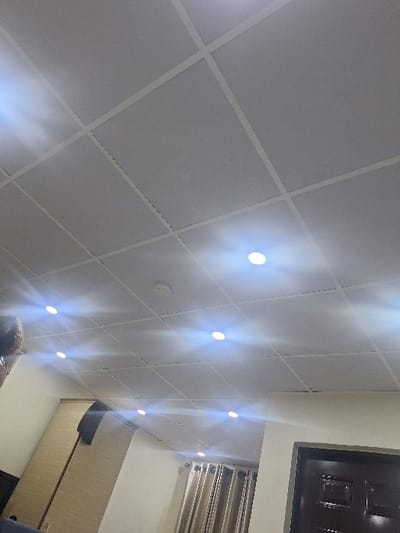 An interior scene in Close, Nigeria, features a bright room. The ceiling is a light gray grid-panel drop ceiling, illuminated by at least seven recessed circular LED lights that emit noticeable lens flares. A white, circular smoke detector is centrally positioned on the ceiling. On the left, a partial view of an individual's head, characterized by curly dark hair and spectacles, is visible. Below the left side of the ceiling, a tall, light brown wooden wardrobe or cabinet with a dark vertical accent is present, with a dark garment hanging on it. The right side of the room shows an off-white wall, a dark brown paneled door on the lower right, and a window covered by light brown/beige curtains with vertical folds. The overall environment suggests a well-lit, furnished indoor space.