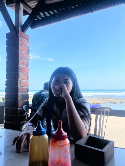 A young woman with long, dark hair is captured in a close-up, sideways shot. She is leaning on her hand, with her face partially obscured by her fingers. She appears to be looking directly at the camera, with a thoughtful or perhaps playful expression. Her attire includes a dark, striped shirt.
The scene appears to be an outdoor dining area, likely at a restaurant or cafe, with a view of the ocean and beach. The background shows a bright, clear sky and the distant coastline with waves. There are also elements of the setting visible, such as a brick wall in the foreground and wooden structures overhead, suggesting a rustic or beachside ambiance.
On the table in front of the woman, there are two plastic squeeze bottles, one containing a red liquid (likely ketchup or chili sauce) and the other a yellow liquid (possibly mustard or a similar condiment). A dark, rectangular object, possibly a napkin holder or a decorative item, is also on the table. A chair is visible in the background, and part of another person's patterned shirt can be seen, indicating the woman is not alone.
The lighting suggests it is daytime, with ample sunlight. The overall mood seems relaxed and casual, possibly a moment of pause during a meal or a break at the beach. The location context indicates this is Pekutatan, Indonesia. There is no visible text in the image.