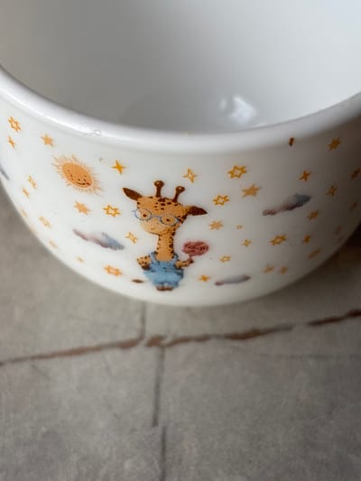 This is a close-up, eye-level shot of a white ceramic mug with a cartoon giraffe design. The giraffe is wearing glasses and blue overalls and is holding a pink object. The mug is adorned with stars and a smiling sun. The background is blurred and appears to be a gray, textured surface, possibly a tabletop or floor, with visible grout lines. The image does not contain any information about the city of Imeni Morozova, Russia.