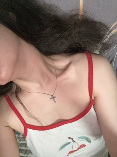 The image is a close-up shot of a person's upper chest, neck, and shoulder, lying down with their head to the left. They are wearing a white tank top with red trim and a cherry pattern. A silver cross necklace is visible around their neck. Dark brown hair is spread across the image. The background appears to be a bed with grey and patterned bedding. The location is not discernible from the image.