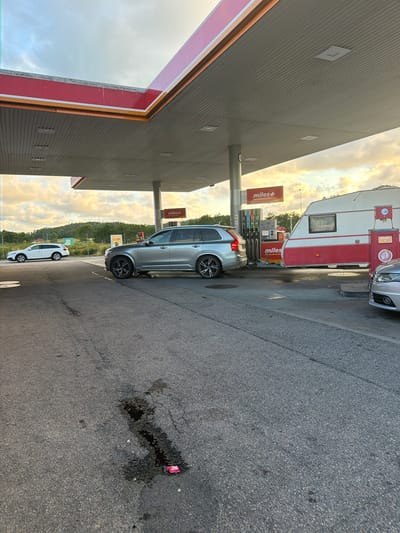 The image depicts a gas station in Hogstorp, Sweden, under a cloudy sky. The foreground is an asphalt-paved area, showing some oil stains and debris, including a red wrapper. A silver Volvo SUV is parked at a gas pump under a red and white canopy. To the right of the Volvo, a white and red caravan is parked, also near a gas pump. Further to the left, a white sedan is visible, parked at a distance. Several "Miles+" signs are present, indicating the gas station brand. In the background, there are trees and hills, suggesting a rural or suburban setting. The lighting indicates it is either early morning or late afternoon. No people are visible in the scene.