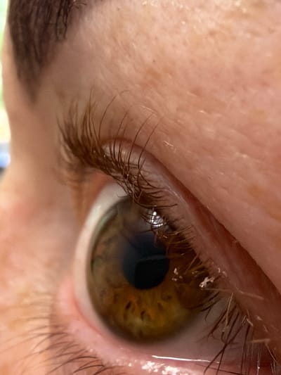A macro photograph captures a close-up view of a human eye. The iris is a striking hazel color with prominent dark brown flecks and a dark pupil. The eyelashes are dark brown and appear somewhat dry, with a few small white specks clinging to them. The skin surrounding the eye is light-toned, showing fine lines and pores, particularly on the eyelid and brow area. The lighting suggests a natural, perhaps indoor, setting, creating subtle highlights and shadows on the skin. The image is cropped tightly, focusing exclusively on the eye and its immediate surroundings, with no background elements visible that would indicate a specific location. The photograph was taken in the city of Узюково, Russia.
