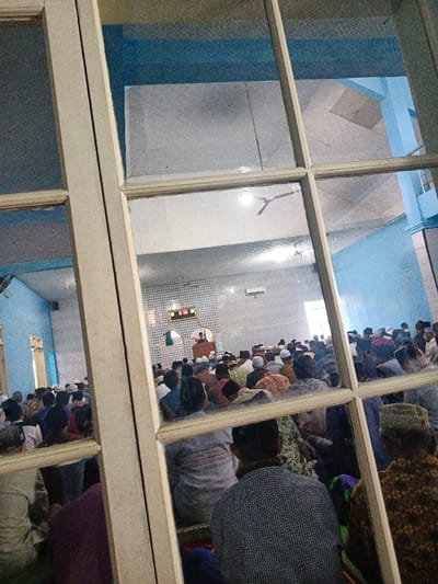 A mosque is seen through a multi-pane window. Inside, many men are seated on the floor, most wearing hats. A man stands at a raised platform near a digital clock and a decorative wall. The room has light blue walls, and ceiling fans are visible. The view is somewhat distorted by the window.