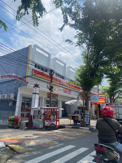 The image shows a street scene in Mojokerto, Indonesia, at the coordinates -7.478 latitude and 112.44 longitude. A large, multi-story building with a red and yellow awning is visible on the left side of the frame. The building is adorned with various signs and banners, including the word "PANGGREMANA" written in large letters.
The street is paved with asphalt and has a crosswalk. A red and white street lamp is positioned at the edge of the sidewalk.
On the right side of the frame, a person wearing a red helmet is riding a motorcycle. The motorcycle is parked in front of a small building with a red sign.
In the foreground, there are several street vendors selling their wares, including a fruit vendor and a food vendor.
The sky is partly cloudy, and there are power lines running overhead. A large tree with green leaves is on the right side of the frame.
