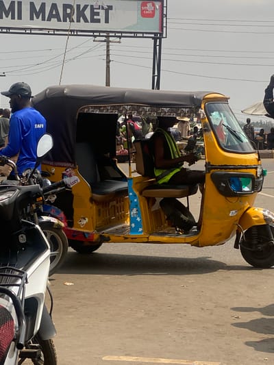 This image depicts a bustling street scene in Ibadan, Nigeria, specifically near a location identified by an overhead sign as "OMI MARKET."
The central subject is a yellow, three-wheeled commercial tricycle, commonly known as a Keke Napep, positioned on an asphalt road. The tricycle has a black canvas roof trimmed with a repeating "Fendi" style pattern. The driver, a dark-skinned male, is seated inside, wearing a dark sleeveless shirt and a neon green reflective safety vest. He holds a stack of banknotes in his left hand. The tricycle's body is adorned with various stickers, including a stylized female face with "Wizzy" text, blue panels with white stars, and patches of red, blue, and leopard-print paint, indicating personalization and wear.
To the immediate left and slightly forward of the tricycle, a dark-skinned male sits on a motorcycle, facing away from the viewer. He wears a black baseball cap and a royal blue long-sleeved shirt with the word "STAFF" printed in white on the back. Partially visible in the lower left foreground is the front section of a white and black motorcycle, featuring a black cargo rack and a red and black patterned seat cover.
In the background, a busy market environment is evident, populated by numerous individuals, some appearing to be vendors. One person wears a distinctive straw hat. Market stalls and an umbrella are visible in the middle distance. Overhead, a large white billboard sign with "OMI MARKET" in black capital letters spans the upper frame, alongside a smaller "SM cash" sign. Utility wires crisscross the sky, connecting to a wooden utility pole. The sky is bright and appears overcast or hazy. The road surface is asphalt, showing minor debris and shadows cast by the vehicles.