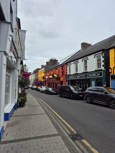 The image depicts a street scene in Carrick-on-Shannon, Ireland, characterized by a row of colorful buildings and parked cars. On the left, a white building with a blue base features large windows, and a sign with red lettering, possibly indicating a business. Adjacent to it, potted plants with pink and white flowers add a touch of greenery. The street itself is paved with grey cobblestones on the sidewalk and asphalt on the road, marked with yellow lines.
Several cars are parked along the street, including black sedans, a black van, and a grey SUV. Across the street, a line of buildings presents a vibrant facade, painted in various colors such as red, yellow, and white with black accents. One prominent building displays a sign that reads "CARRICK BAPTIST CHURCH." Flags and more flowers adorn the storefronts. The sky is overcast with grey clouds.