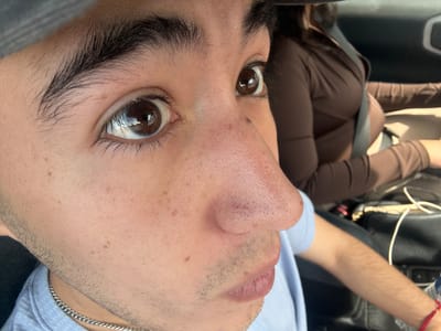 The image is a close-up, fisheye-lens photograph of a young person's face, with a focus on their eyes, nose, and mouth. The person is wearing a light blue collared shirt and a silver chain necklace. Their skin has visible pores and some light freckling. They have dark, thick eyebrows and dark brown eyes that reflect light. The background is blurred, but it appears to be the interior of a car, with another person in the passenger seat wearing a brown top and a seatbelt. A black bag and a water bottle are also visible in the background. The photograph was taken from a low angle, looking up at the subject. There are no identifiable landmarks or indicators in the image to suggest it was taken in Monterrey, Mexico.