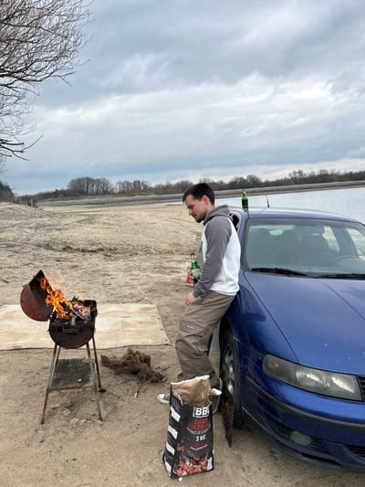 A single adult male is depicted standing on a sandy embankment near a body of water in Любен, Bulgaria, under an overcast sky. The male, appearing approximately 25-35 years old with short dark hair and stubble, is wearing a two-tone grey and white hoodie, khaki cargo pants, and white sneakers. He is leaning against the front passenger side fender of a dark blue sedan, looking downwards and to his left. He holds a green bottle in his left hand. Another green bottle is visible resting on the car's roof, near its antenna.
To the left of the male and car, a rusty, drum-style barbecue grill with its lid open displays active orange flames and burning charcoal. A light-colored rectangular mat or pallet lies on the ground between the grill and the car. Adjacent to the car's front wheel, an open black and white bag labeled "BBQ GRILL HOLZKOHLE BARBECUE CHARCOAL 3 KG" stands upright. Several other green bottles are visible on the ground near the car. The immediate ground consists of light brown sand or dirt, with sparse, bare tree branches visible in the upper left corner. In the mid-ground, a calm body of water extends horizontally, bordered by a line of bare trees and vegetation under a heavily clouded, grey sky.