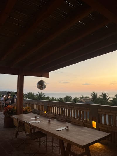 A horizontal, medium-distance shot depicts an outdoor setting with a table, chairs, and an ocean view at dusk in Brisas de Zicatela, Mexico.
To the left, a wooden structure provides a roof over a portion of the scene. Hanging from the structure is a spherical, mirrored object.
In the distance, an ocean and a strip of beach are visible under a yellow and orange sky. Palm trees line the beach.
To the right, a long wooden table with several chairs is set up, with two glasses and napkins on it. Behind the table, a low brick wall with window-like openings runs horizontally. A warm light emanates from one of the openings. At the edge of the frame, there are visible stone-like tiles.
At the bottom of the frame, several people stand near the edge of the structure, with some carrying objects and appearing to be interacting. A potted plant is visible at the bottom.