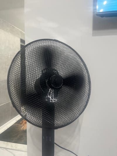 A black standing fan is positioned in front of a white wall. The fan's blades are blurred, indicating it is in operation. To the left of the fan, a tiled wall is visible. In the lower left portion of the image, a portion of a mat with an image of a lion's mane and paws can be seen. The scene appears to be indoors, possibly in a domestic setting within the city of Rosenovo, Bulgaria.