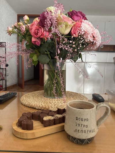 The scene depicts an indoor still life, located in Miljevci, Montenegro. A prominent floral bouquet, comprised of various pink, white, and maroon roses, light pink carnations, and delicate pink filler flowers, stands in a clear glass cylindrical vase, approximately half-filled with water. The vase rests centrally on a circular woven placemat on a glossy, light-brown wooden table. To the left of the bouquet, a black multi-button remote control is present. To the right, a light-colored, speckled ceramic mug with a handle contains a dark beverage and features Cyrillic text: "ДОБРО ЈУТРО ЛЕПОТИЦЕ" (Good Morning, Beautiful), along with a patterned black and white base. Adjacent to the mug, an oval bamboo tray holds approximately 15-18 heart-shaped chocolates, some plain and some topped with a light granular substance. A second black remote control is partially visible behind the mug, and a clear plastic container with yellow/white contents is at the far right edge of the table. The background reveals a light-colored wall to the left, an open wooden doorway leading into another room with a tiled floor, and white kitchen cabinets or furniture on the right. No individuals are present, and no active interactions are depicted.