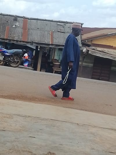 The image shows a full-body shot of an older man walking on a paved road in Otulu, Nigeria. He is wearing a dark blue robe and a patterned cap. In his right hand, he holds a white string of prayer beads. He is also wearing red open-toed sandals. To the left of the man, a blue motorcycle is parked. Behind the man, a row of buildings with corrugated metal roofs is visible, suggesting a market or commercial area. The buildings appear weathered, with some signs of disrepair. In the background, other people and objects are indistinct. The sky is overcast, indicating it might be daytime with cloudy weather. The overall scene depicts a typical street view in a Nigerian town.