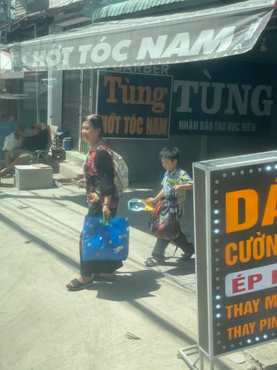 The image depicts a street scene in Phường An Tịnh, Vietnam. In the foreground, an elderly woman in a floral shirt and black pants walks from left to right, carrying a large blue shopping bag. To her right, a young boy in a checkered shirt and dark pants walks in the same direction, carrying a backpack and what appears to be a toy. Behind them, a sign with Vietnamese text is visible, with the words "HOT TOC NAM" and "TUING TUNG" prominently displayed. Further in the background, a person is seated in a chair outside a building, partially obscured by a white cooler. The environment appears to be a commercial street with shops and awnings, under a bright, sunny sky casting shadows on the pavement.
