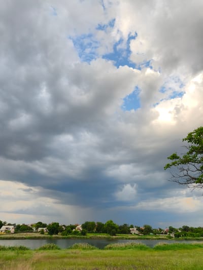 The image depicts a landscape scene in Shyroka Dacha, Ukraine. The foreground features tall, dry grass and a body of water, likely a river, separating the viewer from a village. The village is situated on the opposite bank of the water and is characterized by houses with pitched roofs, interspersed with numerous green trees. A notable feature is the dramatic sky, filled with a mix of dark, heavy storm clouds and patches of bright blue sky. In the distance, a subtle rain shower is visible falling from the dark clouds over the village. The overall impression is one of a rural setting under an impending or ongoing weather event.