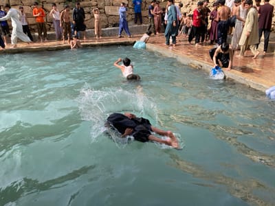 The image captures an outdoor recreational scene at a man-made pool in Killi Malik Sher Mohammad, Pakistan, under direct daylight. Approximately two dozen males, predominantly youths and young adults, are gathered around and within a rectangular pool of light blue-green water. In the immediate foreground, one individual in dark clothing is submerged face-down, generating a substantial splash, with their feet visible extending to the right. To the left, another individual wearing a white sleeveless top is partially submerged in the water. Towards the right side, an individual kneels at the pool's edge, scooping water with a small blue plastic bucket.
Numerous other individuals, dressed in various traditional Pakistani garments, are standing on the reddish-brown paved perimeter surrounding the pool. Some individuals on the edge have their clothing rolled up or appear prepared to enter the water. In the background, behind the assembled people, a dry-stacked stone wall is visible, indicating a rugged, arid environment. The overall scene depicts a communal gathering and recreational activity in the water.