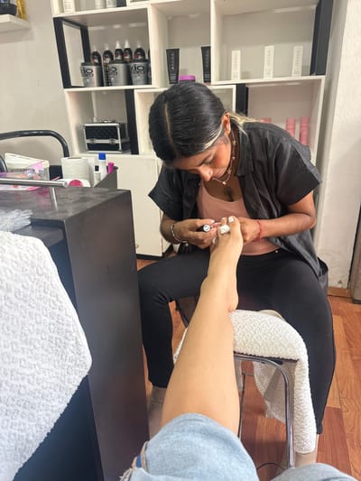 A woman in black leggings and a black smock paints a client's toenails white in what appears to be a nail salon. The woman doing the pedicure has dark hair with a blonde streak and is wearing a necklace. A white towel is placed on the footrest. Nail products and tools are visible on shelves and on a black counter. The location is indicated as San Pedro Cholula, Mexico.