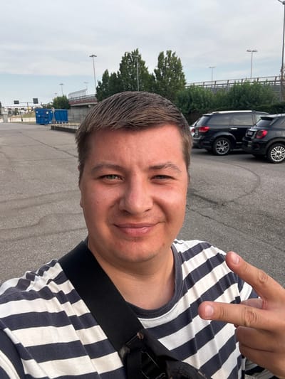 A man is taking a selfie outdoors in a parking lot. He is wearing a horizontally striped t-shirt and has a black strap across his chest, possibly from a bag. His right hand is raised with his index and middle fingers extended in a peace sign. In the background, there are several parked black SUVs and a blue shipping container. Further back, a bridge and trees are visible under an overcast sky. The ground is asphalt with white line markings. The scene appears to be in an industrial or commercial area, possibly near transportation infrastructure, consistent with locations found in San Martino Buon Albergo, Italy.