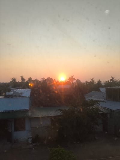 The image presents a wide view of a residential area in Sơn Tịnh, Vietnam, at sunrise or sunset. The sky displays a gradient from light greyish-blue at the top, transitioning to a prominent orange-yellow hue near the horizon. The bright, intense orange-yellow sun is visible near the center of the horizon, partially obscured by a dense line of silhouetted trees, primarily palm varieties, and emits strong light rays and distinct lens flare artifacts. Numerous small, dark, circular specks are scattered across the entire image field. Below the tree line, several buildings are discernible. In the mid-ground, a structure features a dark reddish-brown tiled roof. To its left, a building with a light blue corrugated metal roof and lighter-toned walls is visible, including a dark rectangular opening on its facade and a white cylindrical object (likely a water tank or heater) on its roof. To the right, another structure with a grey corrugated metal roof is partially visible. The immediate foreground consists of dark ground with some low-lying green foliage. There are no individuals or discernible activities depicted.