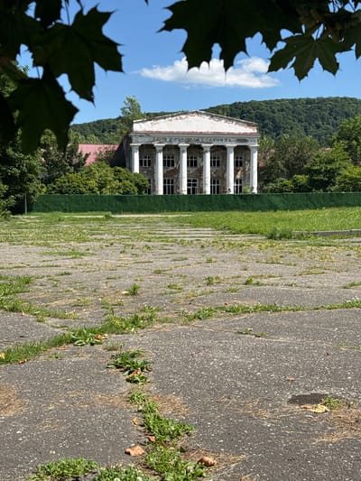 The image depicts an abandoned building with classical architecture, characterized by a white facade and a row of tall columns. The building appears weathered, with peeling paint and darkened window frames, suggesting a state of disrepair and neglect. In the foreground, a cracked asphalt surface is visible, with patches of grass and weeds growing through the cracks, indicating an overgrown and untended environment. To the left of the building, a lush green area of grass stretches out, bordered by trees. In the background, a dense, tree-covered hillside rises under a bright blue sky with scattered white clouds. The scene suggests a warm, sunny day. The overall mood is one of desolation and decay, with nature beginning to reclaim the man-made structure. The leaves of a tree are silhouetted in the left foreground, adding a natural element to the scene. No people or text are visible in the image. The context provided indicates the location is Nalchik, Russia.