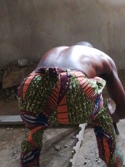 In Enugwu-Ukwu, Nigeria, a shirtless individual is depicted from a rear, slightly elevated perspective, bending forward at the waist. The person, exhibiting a dark skin tone, wears trousers patterned with vibrant green, orange, red, and blue geometric and organic motifs. Their muscular back and shoulders are visible, illuminated by a localized light source creating highlights. The individual's right hand grasps a hammer, identifiable by its dark handle and metallic head. The ground is an uneven surface of dirt and scattered small aggregate. Present on the ground are several pieces of light-colored timber: a long, narrow plank positioned horizontally across the mid-ground, and a smaller, carved piece with an undulating or serrated edge. A portion of a green flip-flop is visible on the individual's left foot, positioned near the bottom right of the frame. The background consists of a plain, light-toned, untextured wall. The scene is consistent with manual labor in a construction or workshop environment.