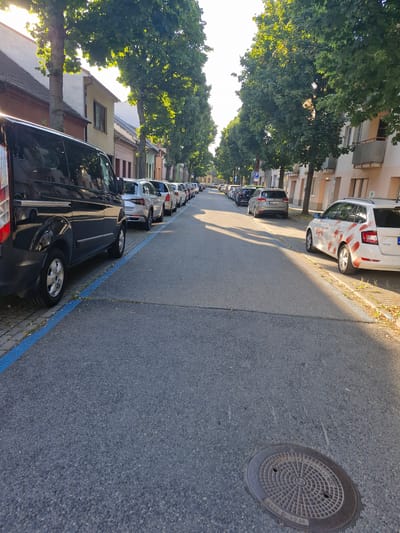 The image depicts a street scene in Brno, Czechia, with a row of cars parked along the side of the road. The perspective is from ground level, looking down the street. Several trees line the sidewalk, their leaves casting dappled shadows on the pavement. The buildings visible are multi-story residential structures with a mix of light-colored facades. A manhole cover is prominent in the foreground on the asphalt. The time of day appears to be late afternoon, with the sun casting long shadows and illuminating the scene with warm light, suggesting clear weather. The parked cars are varied in make and model, with the closest vehicle being a dark-colored van. A blue line is painted along the curb, indicating parking restrictions. There is no visible activity or people in the immediate frame, suggesting a quiet moment. Text is visible on the manhole cover, reading "Kanalizace" and a numerical code. Another piece of text is visible on the white car in the background, potentially a logo or advertisement, but it is too small to be deciphered.