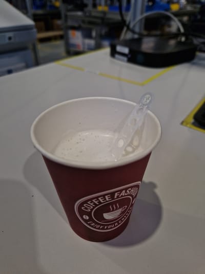 A disposable coffee cup with a logo that reads "COFFEE FASHION ENJOY YOUR COFFEE" is filled with a foamy beverage and has a clear plastic stirrer inside. The cup is red and sits on a light-colored surface, possibly a desk or table, in what appears to be an industrial or office environment with blurred background elements of machinery and shelving. No people are visible in the image. The location is specified as Bahovitsa, Bulgaria.