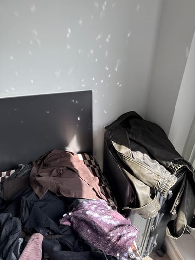 The image shows a cluttered storage space in a location identifiable as Hull, United Kingdom. A black suitcase is partially visible, topped with a dark jacket, suggesting travel or storage of personal items. A pile of clothing in various colors and textures, including a sequined garment, spills out around the suitcase. The storage area is defined by white walls, reflecting dappled sunlight that casts a pattern on the wall and a gray surface, potentially a door or panel.